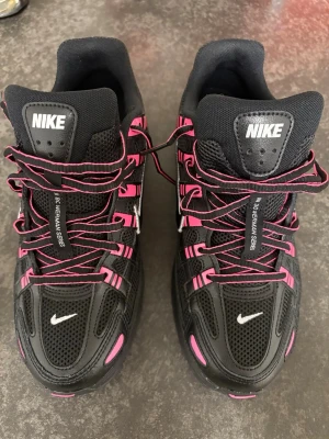 Nike P-6000 svart och rosa sneakers - Nike P-6000 sneakers i svart med rosa detaljer och snörning. Skorna har mesh och syntetmaterial i ovandelen, samt en platt sula och rund tå. Swoosh-logga i silver på sidan och mindre logga på tån. Perfekt för dig som gillar sportig och färgstark stil.