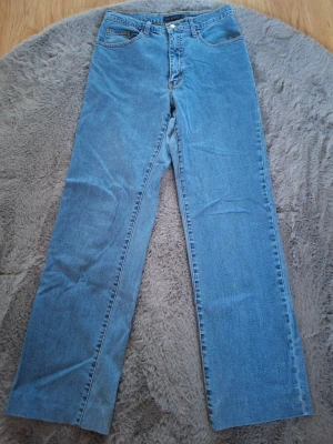 Blå raka jeans från Estelle, strl 36 - Säljer ett par klassiska blå jeans från Estelle i storlek 36. Modellen har raka ben och normal passform, med fem fickor och snygga detaljer på bakfickorna. Jeansen är tillverkade i jeansmaterial och har en tidlös look som funkar till många stilar.