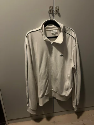 Vit ziptröja från Lacoste - Säljer en vit ziptröja från Lacoste med klassisk ståkrage och svarta detaljer längs axlarna. Tröjan har dragkedja framtill, två fickor och den ikoniska krokodilloggan på bröstet. Perfekt för en sportig och clean look. Storlek Xl men passar L och M ny pris ca 2000 kr helt ny skick använd 1-3 gånger.