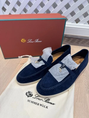 Loro Piana Summer Walk loafers marinblå - Säljer ett par Loro Piana Summer Walk loafers i marinblå mocka med vit sula. Skorna har dekorativt band med metall-detaljer framtill och diskret Loro Piana-logga på hälen. Perfekt för dig som gillar stilrena och lyxiga skor med snygg design. Helt nya, unisex. Låda tillkommer 