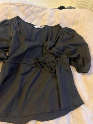 Svart blus med knyt - Säljer en svart blus från 157 med knyt i midjan och puffiga långärmar. Blusen har en djup v-ringning och är tillverkad i ett lätt, ribbat material som ger en snygg struktur. Perfekt att styla med jeans eller kjol.