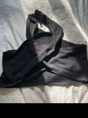 Svart omlott bandeau topp från SHEIN - Säljer en svart bandeau topp från SHEIN i storlek M. Toppen har omlott-design med knytband och är tillverkad i ett vågigt, ribbat syntetmaterial. Perfekt för dig som gillar minimalistisk stil och vill ha en cool topp med öppen rygg.