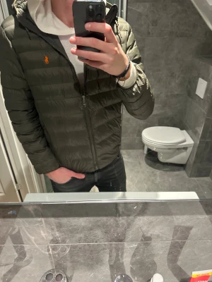 Mörkgrön pufferjacka från Polo Ralph Lauren - Snygg mörkgrön pufferjacka från Polo Ralph Lauren med orange logga på bröstet i riktigt bra skick. Jackan köptes ny för 4000kr. Jag på bilden är 188cm och bär storlek S vilket är lite litet. Hör av er vid funderingar:)