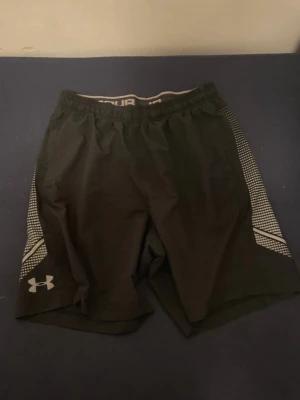 Svarta träningsshorts från Under Armour - Svarta shorts från Under Armour med elastisk midja och snörning. Sidorna har vita prickiga detaljer och loggan i vitt. Modellen är loose fit och materialet är lätt polyester, perfekt för sport och träning.