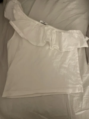 Vit oneshoulder topp med volang - Säljer en vit oneshoulder topp från Kappahl med snygg volang längs halsringningen. Toppen har omlott-design och är ärmlös på ena sidan, vilket ger en cool asymmetrisk look. Perfekt för sommarens alla tillfällen!