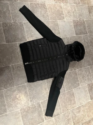 Svart Moncler pufferjacka med huva - Svart cardigan från Moncler med stickade armar samt rygg. Nfc Scan innanför jackan och i loggan på ärmen. Inga defekter. Storlek 3 (M)