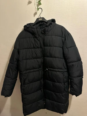 Svart lång pufferjacka från Y.A.S - Svart pufferjacka från Y.A.S i storlek L. Jackan är lång, har huva och dragkedja framtill. Quiltad design med horisontella sömmar och två sidofickor. Perfekt för kalla dagar och har en clean, stilren look.