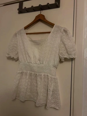 Vit blus med broderade detaljer från SHEIN - Säljer en vit blus från SHEIN i storlek S med broderade hålmönster och puffiga korta ärmar. Blusen har en bred smockad midja och volang nedtill, vilket ger en söt och feminin siluett. Perfekt för vår och sommar, till jeans eller kjol.