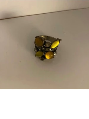 Vacker Rong med stenar  - Unik ring med flera gula stenar i olika former. Ringen har en silverfärgad bas och är dekorerad med små klara stenar som ger extra glans. Perfekt för att lägga till en färgklick till din outfit. Det fattas en Sten men det kan man säkert köpa en passande i butik. 