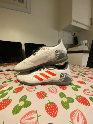 Adidas Copa vita fotbollsskor - Säljer ett par Adidas Copa inomhusskori vitt med grå och röda detaljer. Skorna har klassiska röda Adidas-ränder på sidan, svart snörning och en platt grå sula som passar perfekt för spel på konstgräs eller inomhus. Ovandelen är i syntetmaterial med snygga vågade mönster vid hälen. Storlek 40