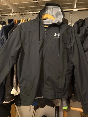 Svart vindjacka från Under Armour - Svart vindjacka från Under Armour med huva och snörning. Jackan har dragkedja framtill, två sidofickor och ett vitt logotryck på bröstet. Lätt och sportig design med meshfoder i huvan, perfekt för aktiva dagar.
