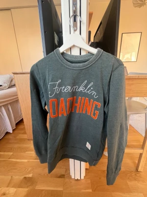 Grön sweatshirt från Franklin & Marshall - Mörkgrön sweatshirt från Franklin & Marshall med trycket 'Franklin Coaching' i vitt och orange framtill. Tröjan har rund halsringning, långa ärmar och mjukt material i bomull. Perfekt för en sportig och avslappnad stil.