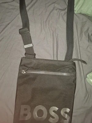 Svart axelväska från BOSS - Säljer en svart axelväska från BOSS med justerbar rem och en stor dragkedja framtill. Väskan är tillverkad i slitstark polyester och har en stilren design med BOSS-loggan tryckt i silver på framsidan. Perfekt för att bära det viktigaste.