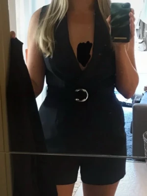 Kostym playsuit med bälte - Snygg mörkblå ärmlös byxdress med djup v-ringning och tillhörande bälte med metallring. Fickor och dragkedja i ryggen. Dressen har en skräddad look och korta ben, perfekt för dig som vill ha en modern och stilren outfit. Materialet känns som polyester och har en slät finish.  Playsuiten är från H&M, lapparna är bortklippta.  Kan tvättas finprogram i maskintvätt 30 grader