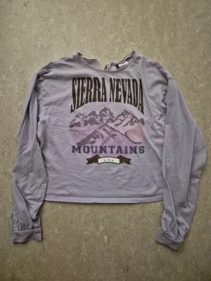 Tröja - Säljer en ljuslila croppad långärmad tröja från H&M med tryck 'Sierra Nevada Mountains' och bergmotiv i lila toner. Tröjan har en avslappnad passform och är gjord i mjuk bomull. Perfekt för chill dagar och snygg till jeans.