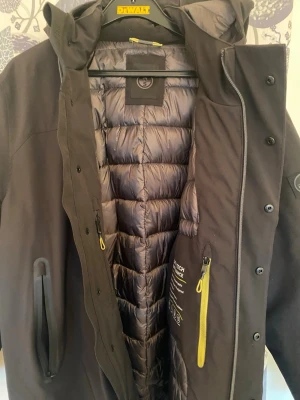 Svart North sail  dunjacka med huva - Säljer en svart dunjacka från North sail med huva och gul dragkedjedetalj. Jackan har återvunnen softshell och termisk isolering, vattentäta dragkedjor och är både vind- och vattenavvisande. Perfekt för kalla dagar och har en modern, quiltad insida. Tvitto finns köpt nyligen använt 2