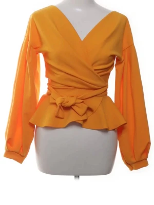 Orange omlottopp med volang och knyt - Snygg gul omlottopp med v-ringning, långa puffiga ärmar och volangkant nedtill. Toppen har ett brett midjeband som knyts i en rosett i sidan för extra detalj. Perfekt för dig som vill sticka ut med färg och form.
