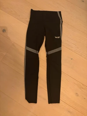 Svarta leggings från Aim'n XS - Svarta leggings från Aim'n i storlek XS med vita prickdetaljer längs benen. Tillverkade i stretchigt material med 73% polyester och 27% spandex, perfekt för träning. Hög midja och smal passform som sitter snyggt och bekvämt.