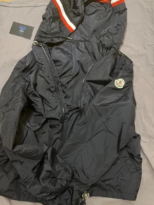 Svart vindjacka från Moncler - Svart vindjacka från Moncler med huva och dragkedja. Jackan har Moncler-logga på ärmen och snygga rödvita ränder på huvan. Perfekt för dig som vill ha en stilren och sportig look. Justerbar med snörning vid huvan.