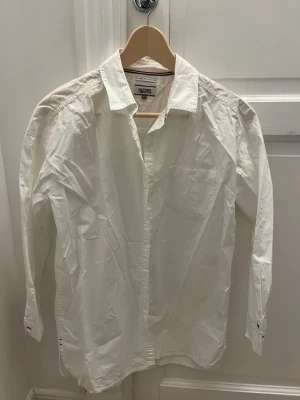 Vit skjorta från Hilfiger Denim XS - Säljer vit skjorta från Tommy Hilfiger Denim i storlek XS. Skjortan har 3-kvartsärm, knappar framtill och en bröstficka. Snygga detaljer med röd och blå söm vid kragen och ärmslut. .
