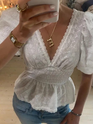 Vit blus med spets och puffärm - Supersöt vit blus med broderade blommor, v-ringning och spetskant. Puffiga korta ärmar och smockad midja som ger en fin siluett. Tillverkad i tunn bomull med volang nedtill. Perfekt till jeans eller kjol för en fräsch look. Man ser vilket möster det är tydligare på sista bilden!