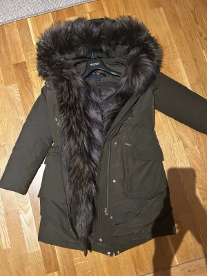 Woolrich dunjacka  - Mörkbrun Whoolrich jacka i nytt skick. Nypris 16.000 kronor. En unik modell med päls hela vägen intill, samt stor och fluffig päls baktill kragen. Jackan har guldiga diskret dragkedja och detaljer. Snygg figurnära passform. 
