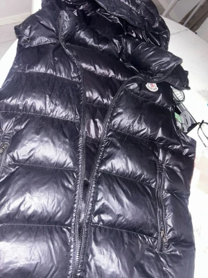 Svart dunväst från Moncler - Snygg svart dunväst från Moncler med glansig finish och klassisk logga på bröstet. Västen har dragkedja framtill, två fickor med dragkedja och hög krage. Perfekt för lager-på-lager och streetstyle.