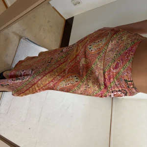 Mönstrad långkjol i paisley - Säljer en färgstark långkjol med paisleyinspirerat mönster i orange, rött, grönt och gult. Kjolen har omlott-detalj i midjan och är tillverkad i ett mjukt, glansigt material som ger ett snyggt fall. Perfekt för dig som vill sticka ut med en bohemisk vibe.