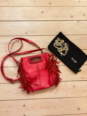 Röd axelväska med fransar från Maje - Cool röd axelväska från Maje i läder med fransar på sidorna och justerbar axelrem. Väskan har ett handtag upptill och kommer med en guldfärgad kedja samt dustbag.