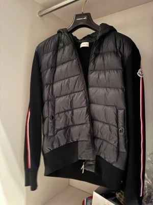 Svart vindjacka från Moncler - Snygg svart vindjacka från Moncler med quiltad front och huva. Jackan har dragkedja, två fickor med knapp och Moncler-logga på ärmen. Ärmarna är svarta med röd, vit och blå rand längs sidan. Perfekt för dig som vill ha en sportig och stilren look. Nypris på Moncler butiken i Stockholm var 10999 när jag köpte den