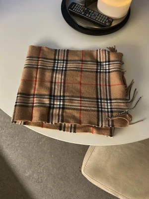 Beige rutig halsduk med fransar - Klassisk beige halsduk med rutigt mönster i svart, vitt och rött. Halsduken har tunna fransar i ändarna och är tillverkad i ett mjukt tyg som känns skönt mot huden. Perfekt accessoar för att lyfta din outfit.