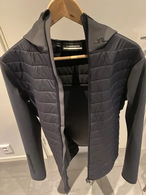 Mörkgrå vindjacka från J.Lindeberg - Snygg mörkgrå vindjacka från J.Lindeberg med quiltade partier framtill och på axlarna. Jackan har hel dragkedja, hög krage och är tillverkad i ett lätt, vindtätt material med stretchiga ärmar för extra komfort. Storleken är S som du ser på bild 4. Pris är diskuterbart.