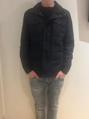 Bondelid field Jacket - Snygg mörkblå vindjacka med fyra stora fickor framtill och knappstängning. Jackan har hög krage och en rak passform som ger en clean look. Perfekt för blåsiga dagar och enkel att matcha med olika outfits. Materialet känns lätt och smidigt.