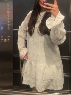 Vit blommig kortklänning med volang - Söt vit kortklänning med blommigt mönster och volangdetaljer. Klänningen har långa, lätt puffade ärmar och breda manschetter. Perfekt för sommarens alla tillfällen och ger en romantisk vibe.