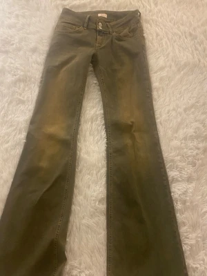 Bootcut jeans i sandfärg - Säljer ett par sandfärgade bootcut jeans med låg midja och klassiska fem fickor. Jeansen har snygga slitningar framtill och bakfickor med lock och knapp. Perfekta för dig som gillar retrovibbar och vill ha ett par unika jeans i garderoben.
