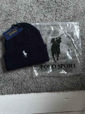 Mörkblå ribbstickad mössa Polo Ralph Lauren - Snygg mörkblå ribbstickad mössa från Polo Ralph Lauren med klassisk broderad logga framtill. Mössan är tillverkad i mjukt material och har en uppvikt kant för extra stilpoäng. Perfekt accessoar för kalla dagar.