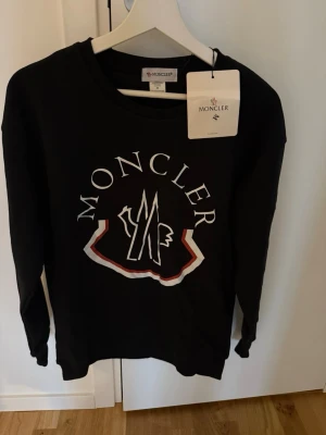 Svart sweatshirt från Moncler - Snygg svart sweatshirt från Moncler med stort vitt och rött logotryck på bröstet. Tröjan har rund halsringning och långa ärmar. Tillverkad i mjukt bomullsmaterial för en bekväm och avslappnad stil.