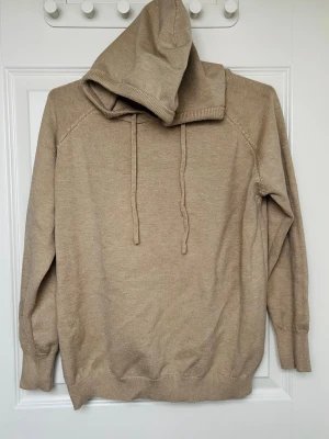 Beige stickad hoodie med huva - Mysig beige stickad hoodie med dragsko i huvan och ribbade muddar vid ärmslut och nederkant. Perfekt för dig som gillar en enkel och stilren look. Materialet känns mjukt och skönt mot huden och passformen är avslappnad.
