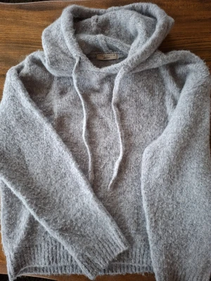 Ljusgrå fluffig hoodie från Gina Tricot - Mysig ljusgrå stickad hoodie från Gina Tricot i fluffigt material. Hoodien har huva med dragsko, långa ärmar och ribbade muddar vid ärmslut och nederkant. 