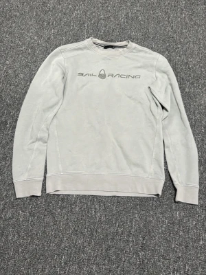 Ljusgrå sweatshirt från Sail Racing - Ljusgrå sweatshirt från Sail Racing med rund halsringning och tryckt logga på bröstet. Tröjan har långa ärmar och ribbade muddar vid ärmslut och nederkant. Perfekt för en avslappnad och stilren look.
