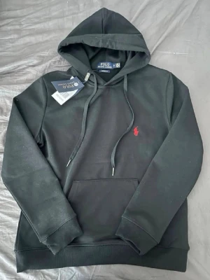 Svart hoodie från Polo Ralph Lauren - Svart hoodie från Polo Ralph Lauren med klassisk huva och dragsnören. Har en känguruficka framtill och den ikoniska röda Polo-loggan broderad på bröstet. Tillverkad i mjukt material som känns skönt mot huden. Perfekt för en avslappnad och stilren look.