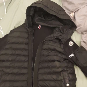 Svart pufferjacka från Moncler - Snygg svart pufferjacka från Moncler med huva och dragkedja framtill. Jackan har quiltad design och ikonisk Moncler-logga på ärmen. Perfekt för kalla dagar och ger en stilren look. Materialet är glansigt och jackan har ribbade detaljer vid ärmarna.