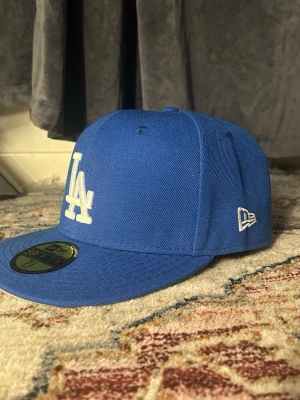Blå LA Dodgers keps från New Era - Säljer en blå snapback-keps från New Era med LA Dodgers-logga broderad i vitt framtill. På sidan finns ett färgglatt broderi med palmer och texten 'Greetings from LA'. Klassisk rak skärm och broderade ventilationshål. Perfekt för dig som gillar streetwear och sportig stil.