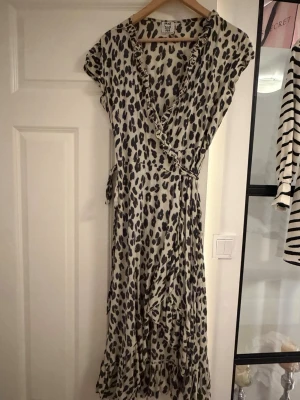 Leopardmönstrad omlottklänning Stajl - Säljer en lång leopardmönstrad omlottklänning från Stajl. Klänningen har volangdetaljer längs kanterna, korta ärmar och knytband i midjan. Färgen är beige med svarta leopardfläckar och materialet känns lätt och luftigt, perfekt för varma dagar.