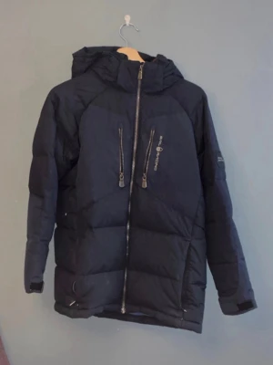 Svart dunjacka med huva från sail racing  - Snygg svart dunjacka från sail racing med huva och två vertikala bröstfickor med dragkedja. Jackan har quiltad design, hel dragkedja framtill och en logga på bröstet. Perfekt för kalla dagar och har en klassisk, sportig look. (Ops! Dragkedjan för att stängas behöver fixas lite)