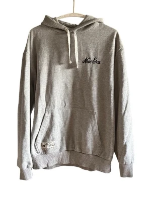 Grå hoodie från New Era med huva - Passar definitivt L också!!! .Grå hoodie från New Era med broderad logga på bröstet och klassisk känguruficka. Huva med vita snören och ribbade muddar vid ärmslut och nederkant. Perfekt för en chill och avslappnad stil.