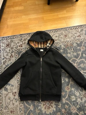 Burberry hoodie - Snygg svart hoodie från Burberry med dragkedja och klassiskt beige-rutigt Burberry-mönster i huvan och längs insidan. Hoodien har en avslappnad passform och är tillverkad i mjukt material. Som helt ny använd fåtal gånger 