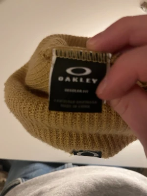 Beige ribbstickad mössa från Oakley - Snygg beige ribbstickad mössa från Oakley med uppvikt kant och diskret logga framtill. Perfekt för att hålla värmen och ge en clean look under kalla dagar. Materialet är mjukt och stretchigt för skön passform.