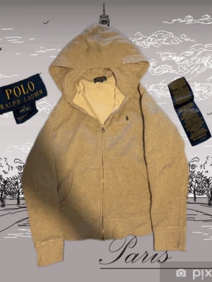Grå hoodie från Polo Ralph Lauren - Snygg grå hoodie från Polo Ralph Lauren med dragkedja och klassisk huva. Diskret broderad logga på bröstet och två fickor framtill. Tillverkad i mjuk bomullsblandning som är skön att bära. Perfekt för en avslappnad och stilren look.strl 158/164