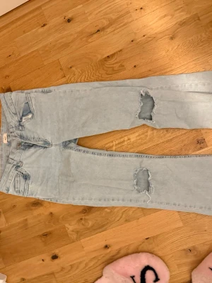 Ljusblå ripped jeans med broderi - Säljer ett par ljusblå jeans med slitningar på båda benen och broderade detaljer på bakfickorna. Jeansen har klassisk femficksmodell och raka ben. Perfekta för en avslappnad och trendig look.